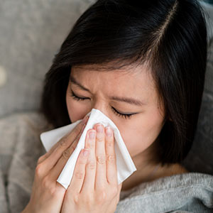 Woman on couch with nasal congestion under blanket blowing nose into tissue